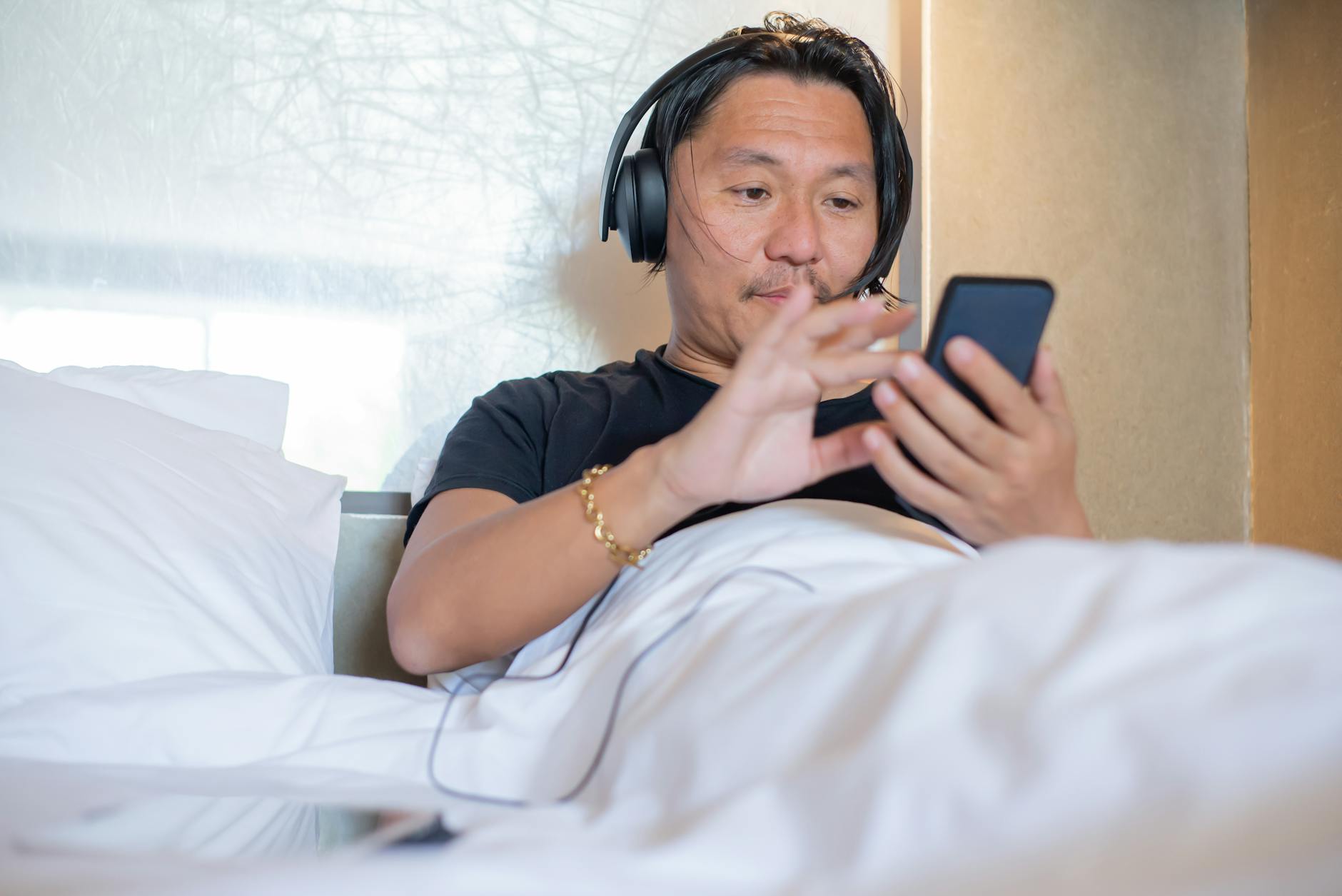 Ranking najlepszych audiobooków klasyki literatury, które dzięki świetnym lektorom zyskują drugie życie Mężczyzna słucha audiobooka w słuchawkach, leżąc w przytulnej sypialni