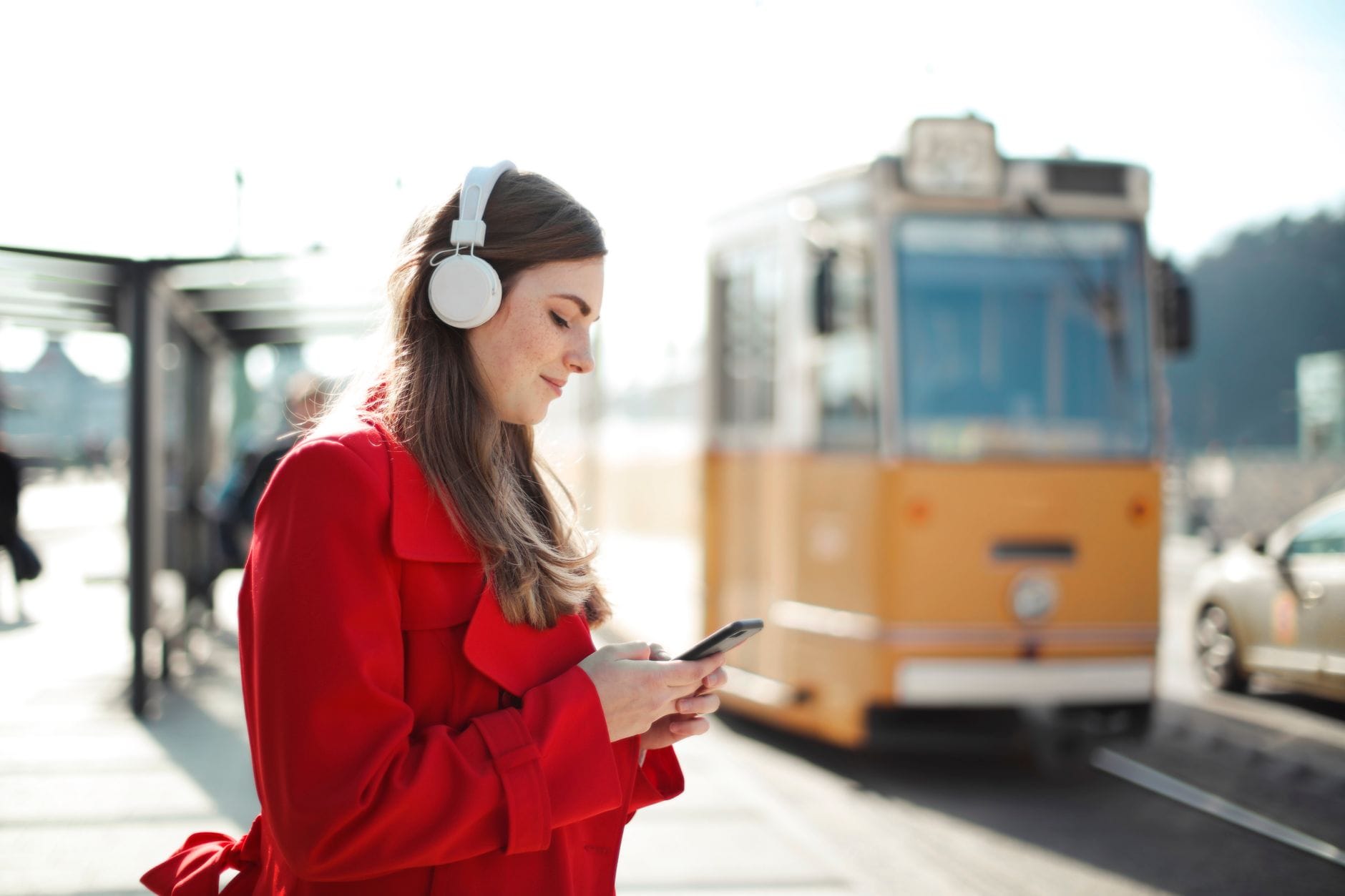 Kobieta w czerwonym płaszczu słucha audiobooka przy przystanku tramwajowym