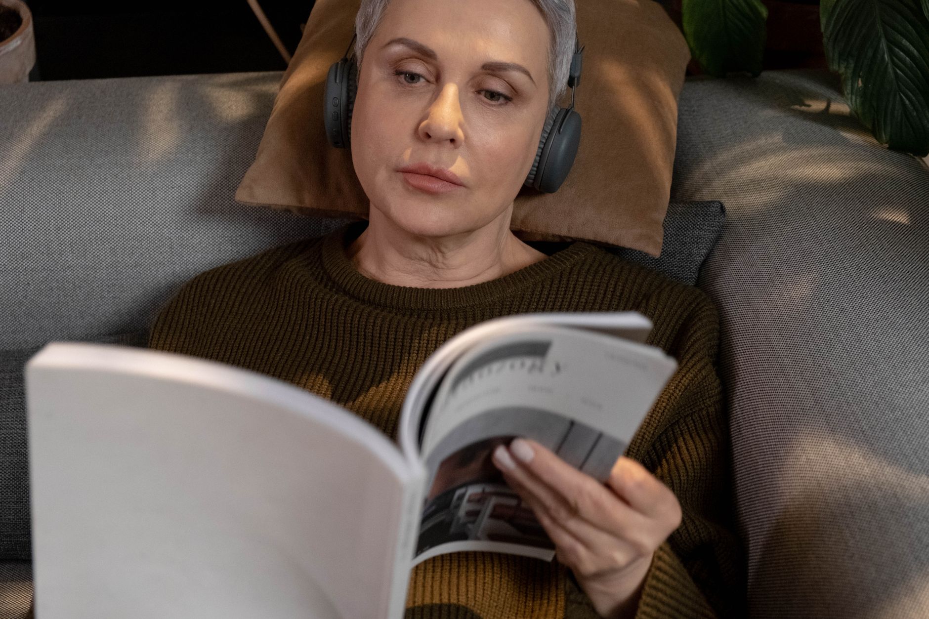 Kobieta w słuchawkach czyta magazyn na kanapie w przytulnym salonie
